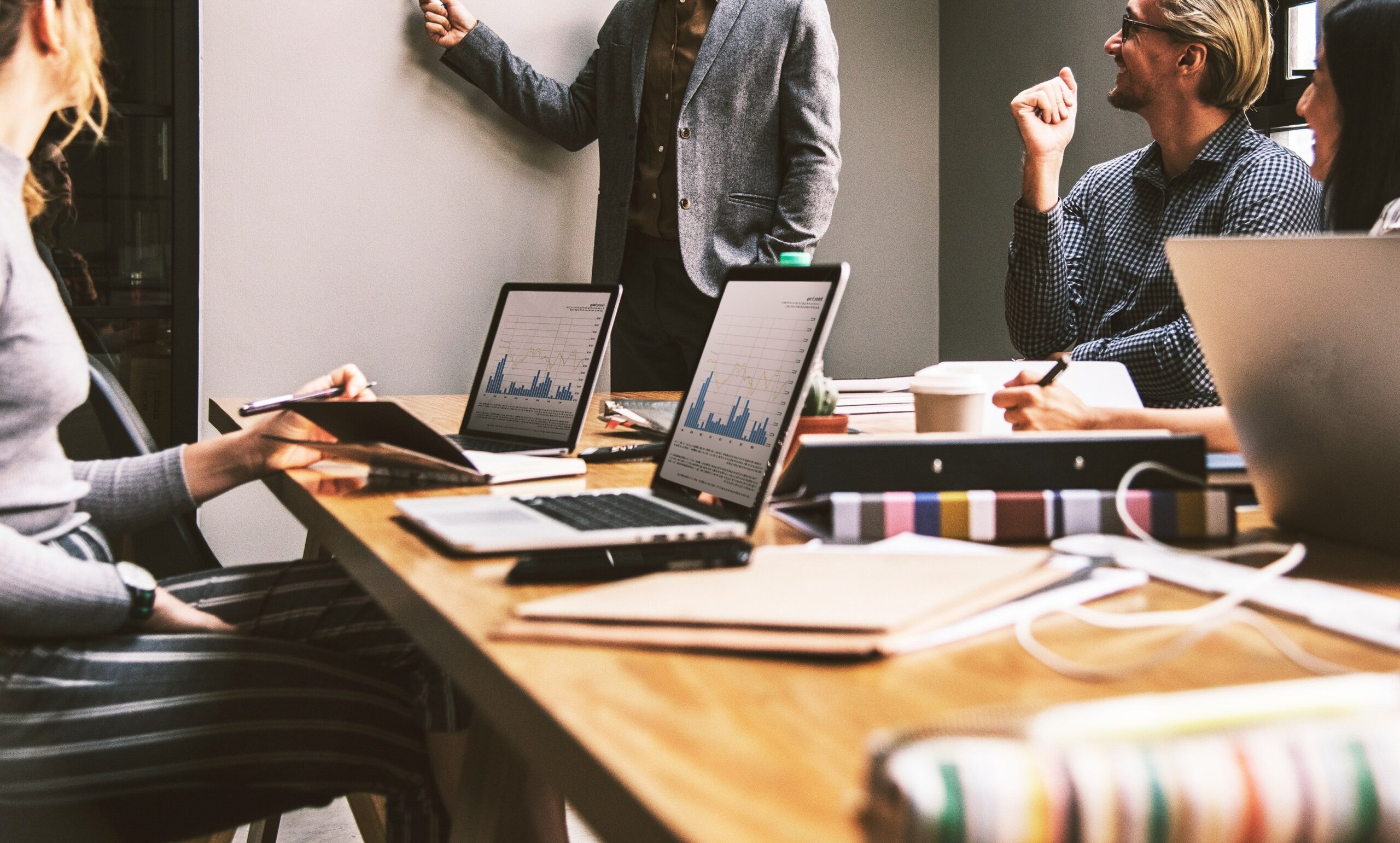 Vier Menschen in einer lockeren, fröhlichen Gruppendiskussion. Drei Personen sitzen, eine davon steht an einem Whiteboard. In der Mitte des Bildes steht ein Holztisch mit Bürounterlagen und 3 Laptops. (Bild von Ronald Carreño auf Pixabay)